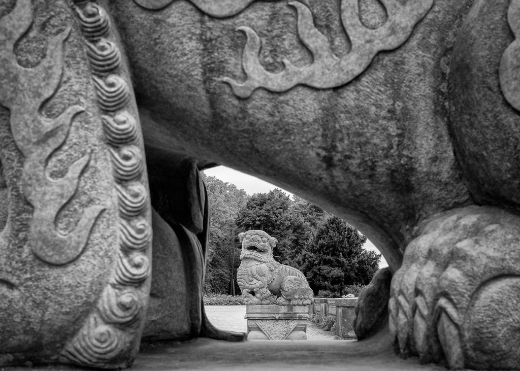 Chinese lion sculpture viewed through the legs of a companion lion sculpture. Highfields Park, Nottingham.