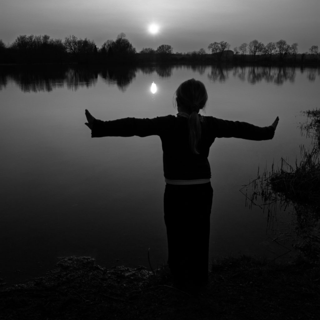 Young girl stood by lake at sunset, arms outstretched.